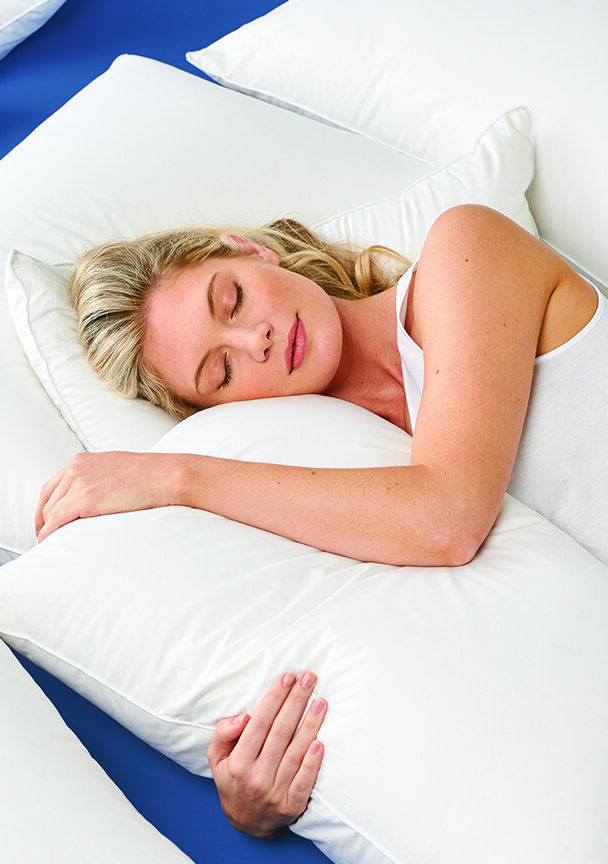 Sleepy blonde model hugging a pillow while laying in a bed of white pillows